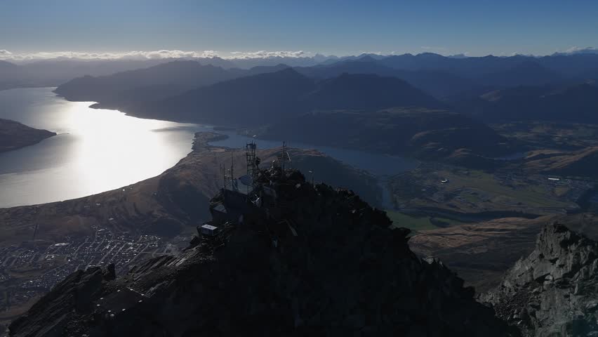 Aerial silhouette of man at comm tower on mtn peak above silvery lake