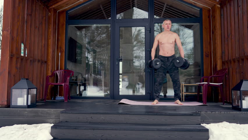 A man does gymnastics with dumbbells on the terrace of his house in winter.