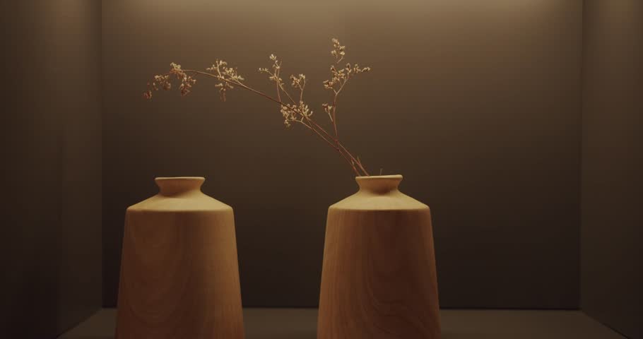 Two empty wooden vases sit on a shelf, illuminated by a warm light. One vase holds a single dried flower stem, adding a touch of simple elegance to the scene.