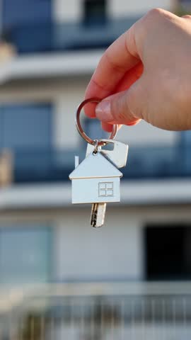 Hand holding house keychain in front of modern apartment building, real estate and homeownership concept.
