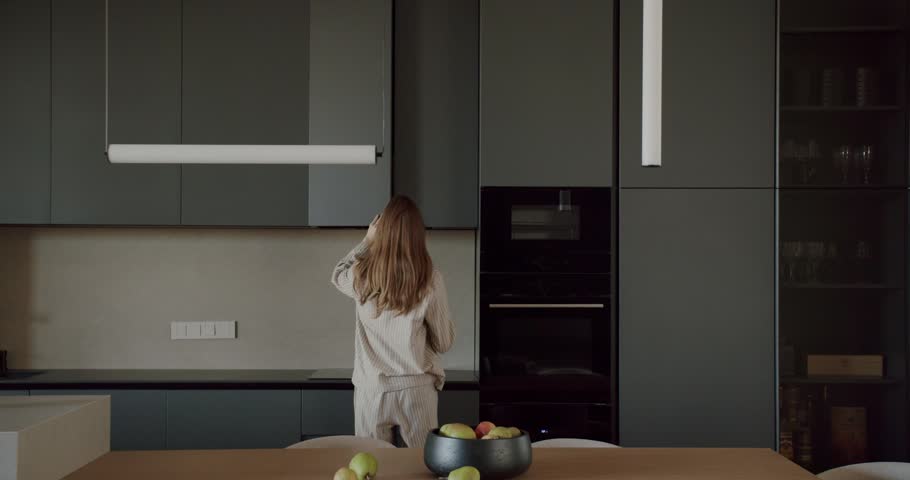 Woman in pajamas opens cupboard in modern kitchen. The contemporary design features dark gray cabinets and sleek appliances. She reaches for something inside, showcasing the practicality and style