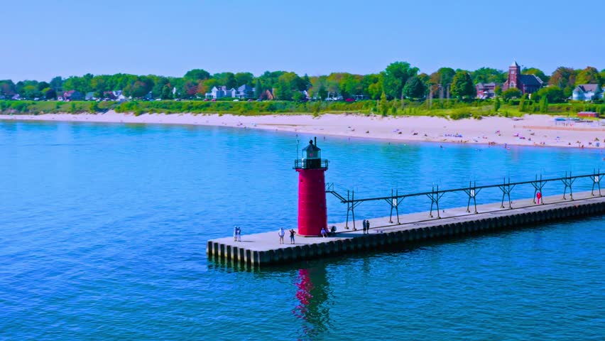 Scenic aerial footage of historic red lighthouse tower standing at end of concrete pier on lake michigan coastline, tourists walking on breakwater walkway during sunny summer day in michigan.