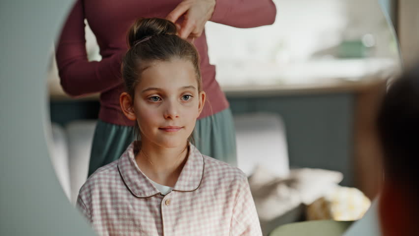 Home kid having hairstyle routine looking mirror closeup. Smiling beautiful mother doing hair bun to daughter morning routine. Happy brunette family admiring reflection in bedroom. Relatives concept 