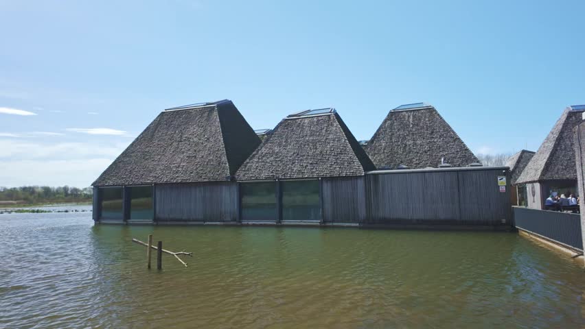 A smooth, detailed pan across the unique wooden-shingled floating visitor village at Brockholes. This footage emphasizes the award-winning sustainable design and geometric architecture of the site, set against a calm lake and clear blue sky in the heart of Lancashire.