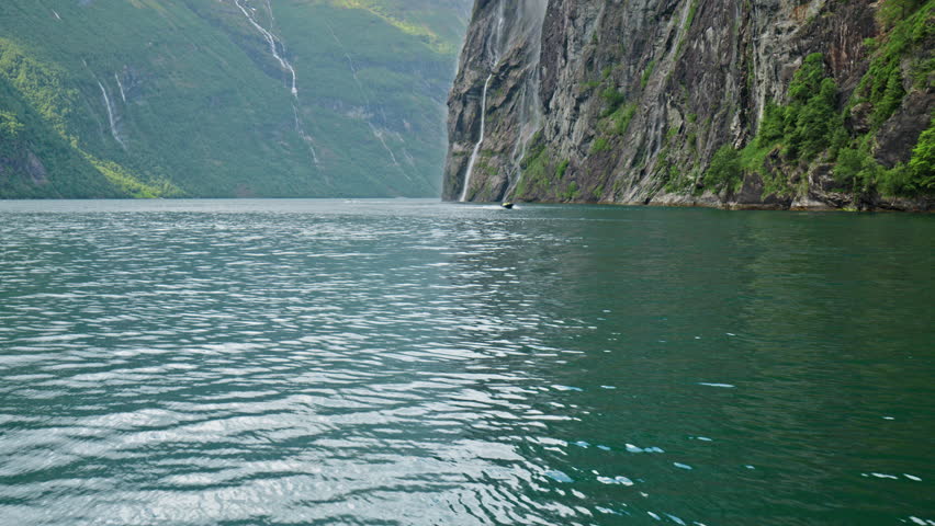 Steep rocky cliff rising directly from calm emerald water in Geirangerfjord. Dramatic fjord landscape.