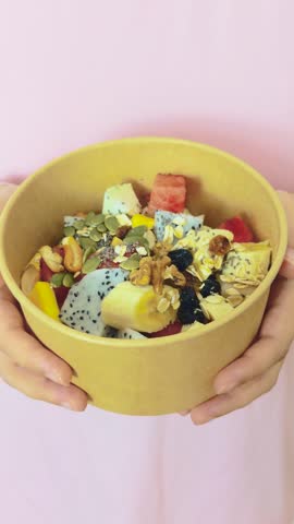 Person holding a vibrant fruit and muesli bowl with dragon fruit, banana, mango, watermelon, seeds and nuts on pink background nutritious, colorful breakfast and snack option