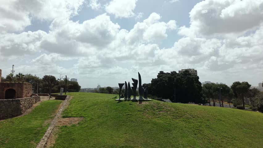 Abstract metal sculptures on green hillside park in jaffa israel, ancient stone wall beside modern art installation under dramatic cloudy sky

