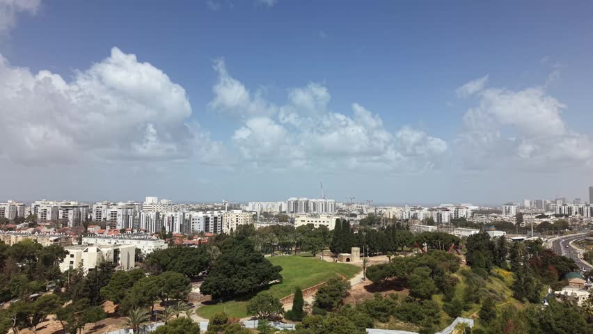 Aerial view of tel aviv skyline with lush urban park in foreground, ancient stone tower amid trees and modern buildings under blue sky

