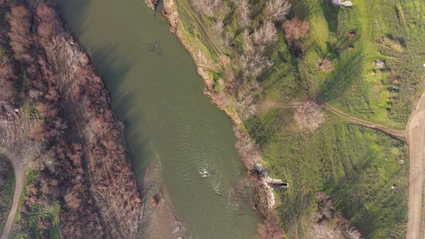 Aerial shots show a river winding through green fields and trees. The landscape features patches of grass and dirt paths near the water. The light of late afternoon casts shadows on the ground. This view highlights the connection between the river and the surrounding environment.