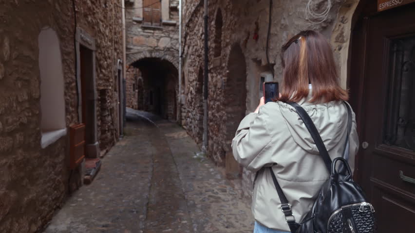 Rear View Of A Young Female Traveler Using Her Smartphone To Capture The Beauty Of A Narrow Stone Alley In A Historic Town. A Modern Travel Experience Within A Traditional Architectural Setting.