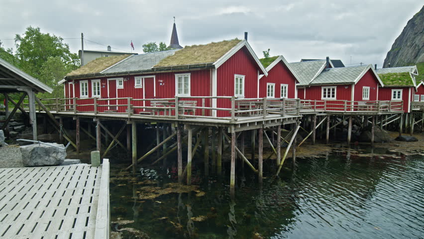 Row of red rorbuer cabins on stilts above water in Lofoten, Norway. Classic fishing village with reflections and mountain backdrop.