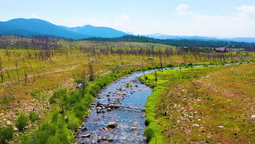 Reverse flying aerial in Rocky Mountain National Park near Grand Lake Colorado revealing a winding stream across open meadow terrain with scattered boulders charred trees and mountain views