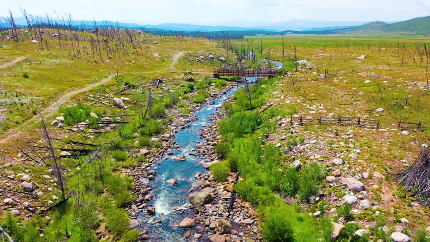 Forward flying aerial near Grand Lake in Rocky Mountain National Park tracking a winding stream past boulders charred trees open meadow terrain and distant Colorado mountain horizons