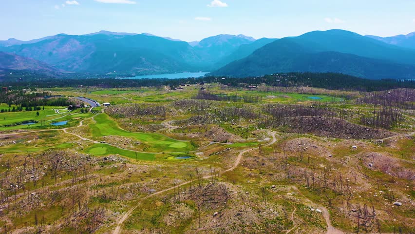 Drone panorama of bright golf fairways carved into a burned mountain landscape, with scorched trees, winding cart paths, and sweeping summer scenery near Rocky Mountain National Park Colorado.