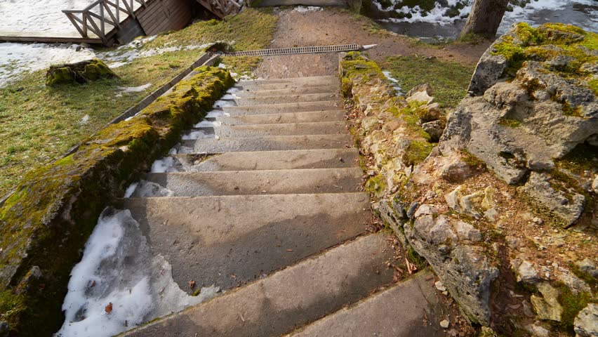 Stone stairs descending beside a crumbling moss-covered wall with loose rubble and traces of snow, showing worn historic masonry, unsafe edges, and early spring thaw.
