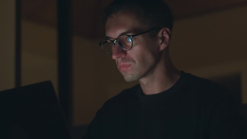 Man working late at night on laptop, illuminated by screen light and fully concentrated. Night work concentration reflects productivity, deadlines, dedication and intense modern workflow