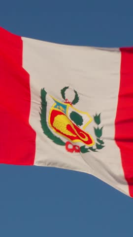 Peruvian national flag proudly waves in the wind against a vibrant clear blue sky. Symbol represents sovereignty and national identity, featuring the coat of arms. Colors of red and white are strik...