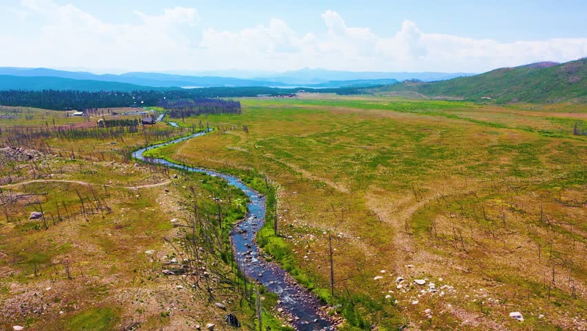 Aerial footage of Rocky Mountain National Park near Grand Lake, Colorado, featuring the broad Kawuneeche Valley, a curving stream, open wetlands, and hazy mountain horizons.