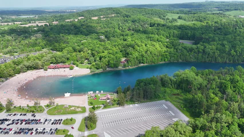 The undulating landscape surrounding a beautiful breathtaking state park, a beach front full of people, and an amazing lake as people enjoy the nice weather.