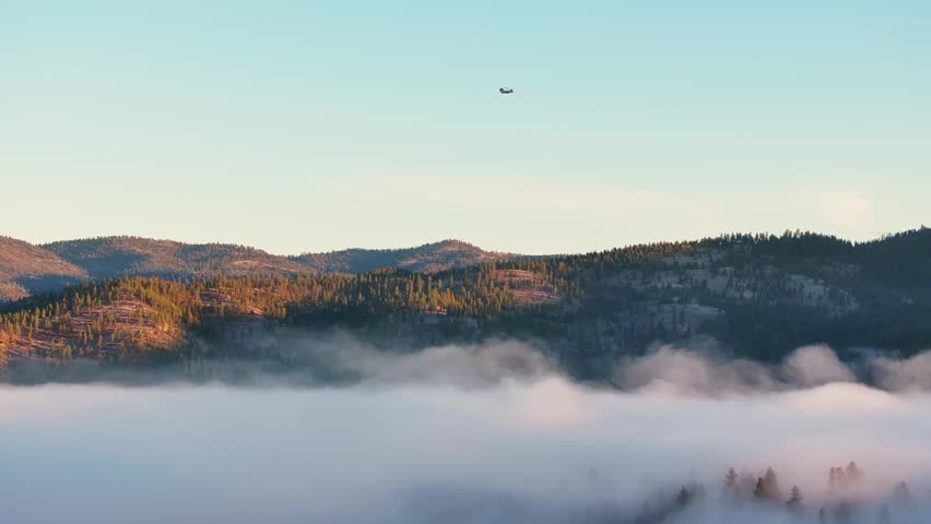 Rolling mountain ridges above low fog and pine forest at sunrise Montana as army transport helicopter flies in sky