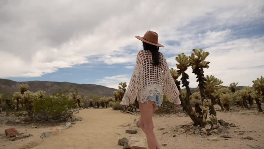 Woman walking along a path through the Cholla Cactus Garden in Joshua Tree National Park, surrounded by dense cactus formations and dry desert terrain. Ideal for travel, lifestyle, and outdoor exploration themes.