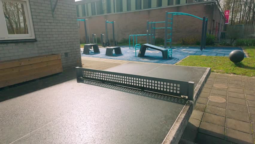 Sunny outdoor sports park featuring a concrete ping-pong table in the foreground and a modern calisthenics gym with blue equipment in the background. Green grass and brick buildings are visible.