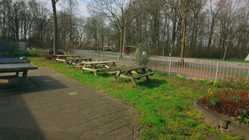 Tranquil outdoor picnic area featuring multiple rustic wooden tables and benches on a lush green lawn. Area is lined with early spring flowers, a paved path, and fence beside road. Bare trees visib...