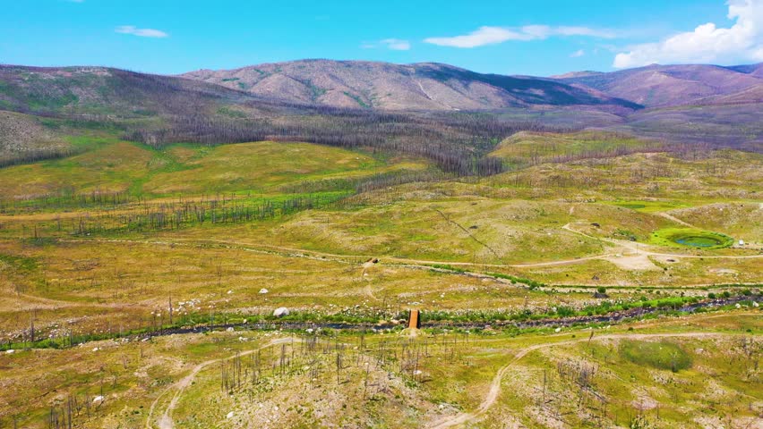 Drone shot of a recovering burn area in Rocky Mountain National Park Colorado, showing charred forest, open meadow, rocky terrain, narrow creek, and mountain slopes in bright daylight.