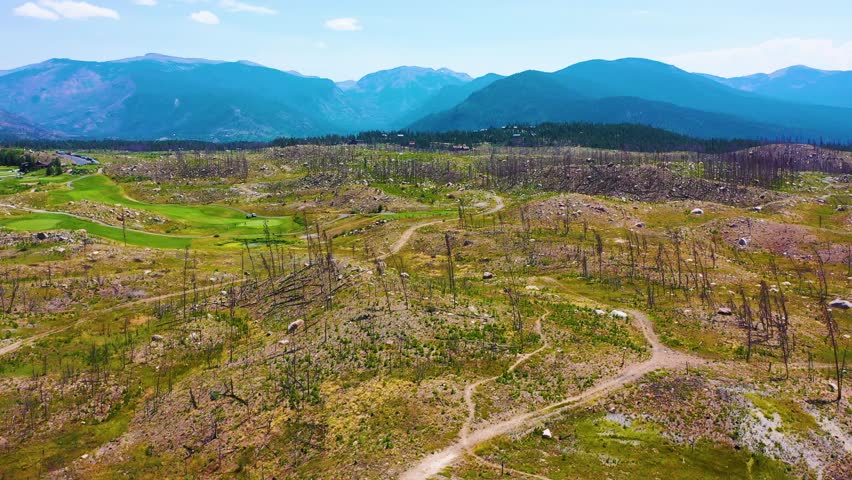 Aerial view of a golf course surrounded by burned forest near Grand Lake Colorado, with green fairways, blue water, scattered cabins, and mountain peaks under a sunny summer sky.