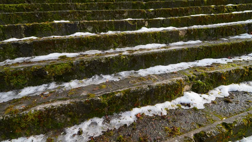 Moss-covered old stone steps with patches of melting snow and ice, showing slippery historic stairway texture, winter wear, and unsafe footing outdoors.