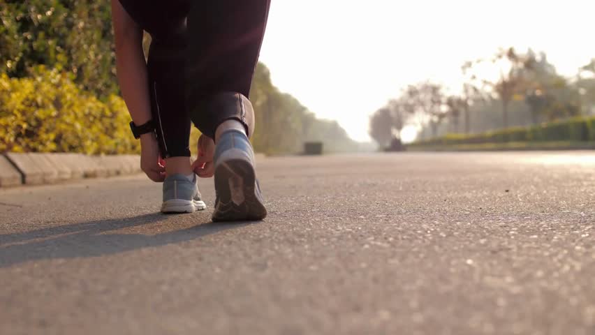 Close up shot of runner woman tying running shoes laces for summer run in public park. Young sporty girl tying shoelace while jogging at public park in the morning. Morning exercise.