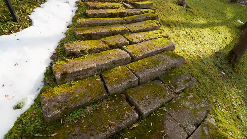 Uneven moss-covered old stone steps on a grassy hillside with melting snow, showing damaged historic access, slippery footing, and erosion in early spring light.