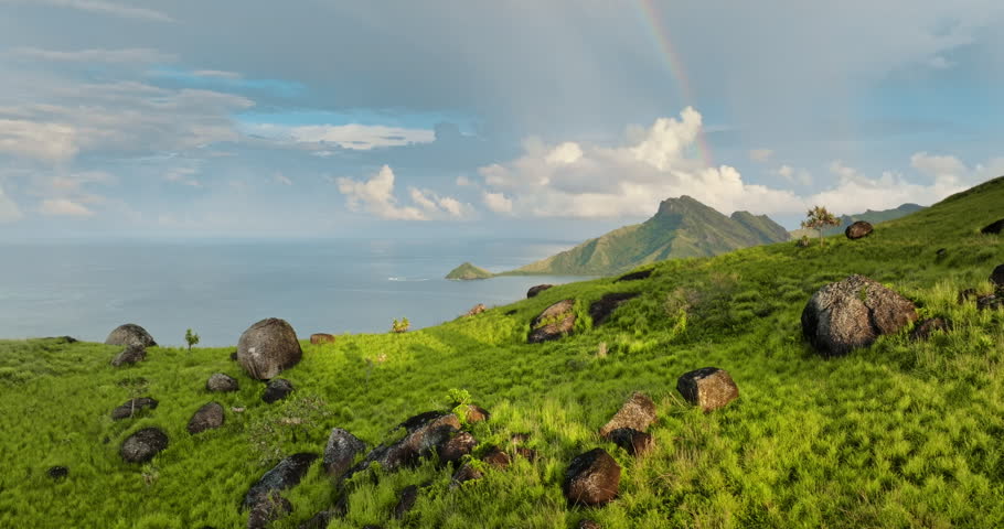 Fiji tropical nature island landscape. Green rolling hills dotted with boulders overlook a tranquil Pacific coastline bay, lush mountains in the distance and a vibrant rainbow in sky. Travel, explore