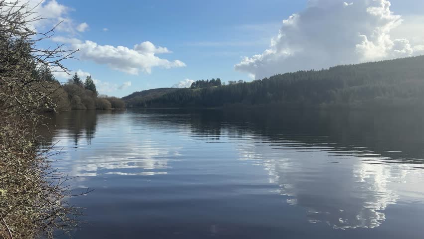 Experience the serene beauty of Brecon Beacons with this tranquil lake scene reflecting forested hills under a bright blue sky. Perfect for peaceful, nature-inspired video projects.