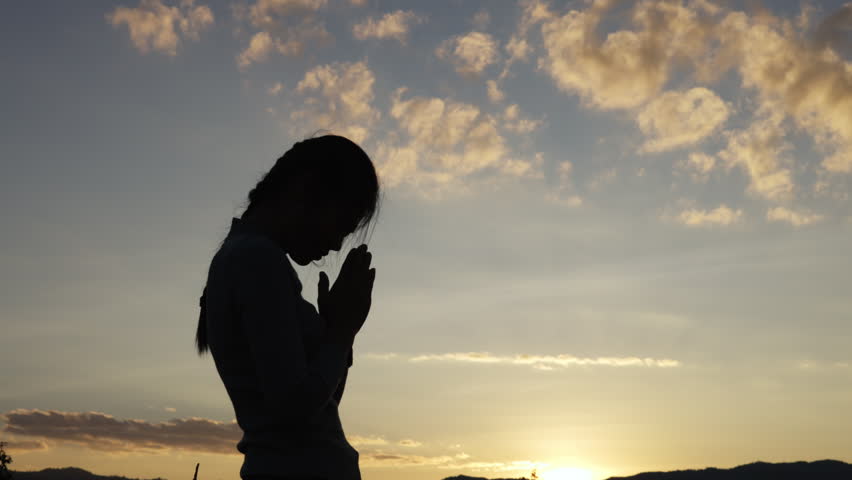 A silhouette of a woman open palm  praying on a mountaintop at sunset, 4K slow-motion video.