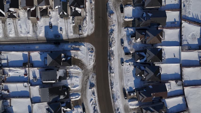 Overhead aerial drone shot of residential street and roundabout in Bridgwater Winnipeg Manitoba. Snow covered houses, yards, and cars on road during a sunny winter day.