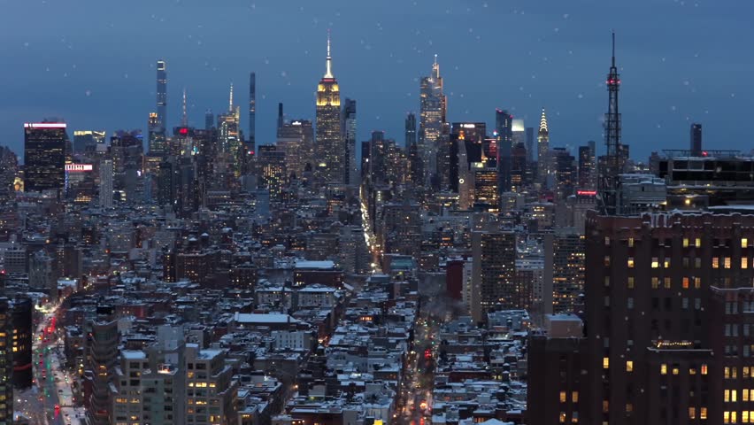 Aerial view of New York City skyline at dusk during snowfall, with Manhattan towers glowing above snow-covered streets and dense urban blocks, capturing cinematic winter atmosphere. Wide shot.