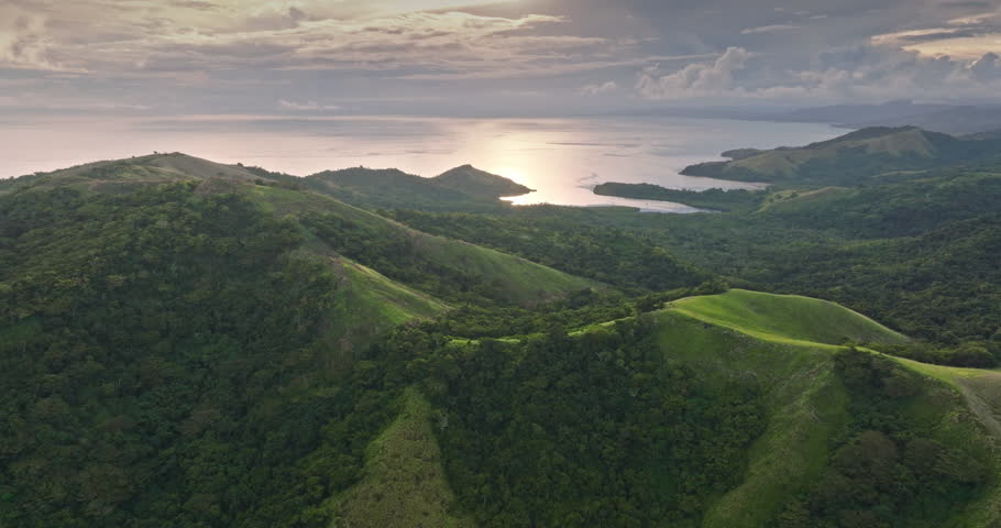 Tropical landscape of Fiji Islands, lush green mountains sloping down to the coastline with the ocean reflecting the late afternoon sun under a cloudy colorful sky. Aerial view drone panorama