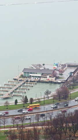 A marina sits by the water with several boats and a city road below on a cloudy day. Trees line the edge of the park.