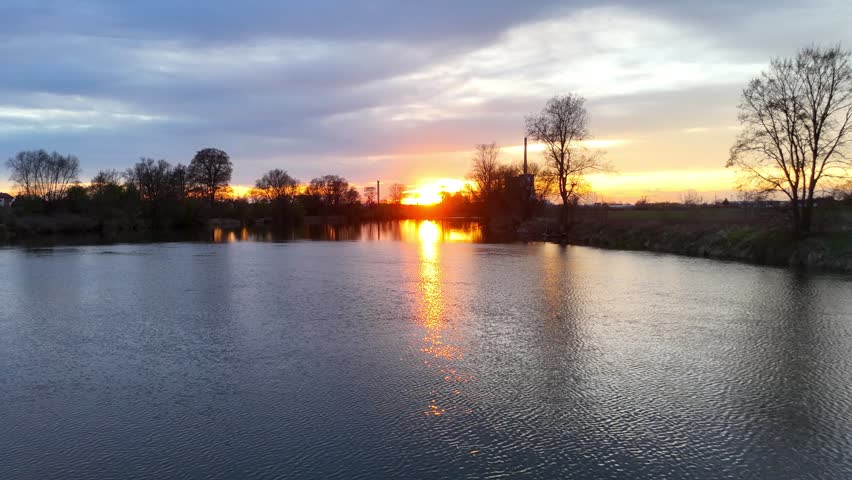 Aerial view of a beautiful sunset over a calm river, showcasing vibrant colors reflecting on the water, with silhouettes of trees and a distant industrial structure in the background