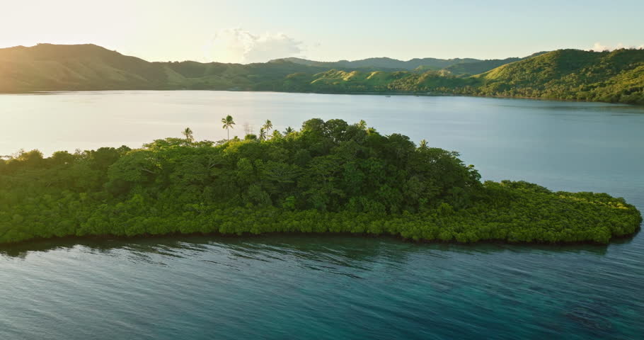 Lush green tropical island bay in Fiji, vibrant mangrove forest, clear turquoise water, and small fishing boat under golden sunset with sun flare and distant hills. Aerial view drone flight panorama