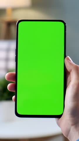 Close up of a modern smartphone with blank green chroma key screen held in one hand against a softly blurred home interior. Mobile device, cellphone or touchscreen display shown vertically for app