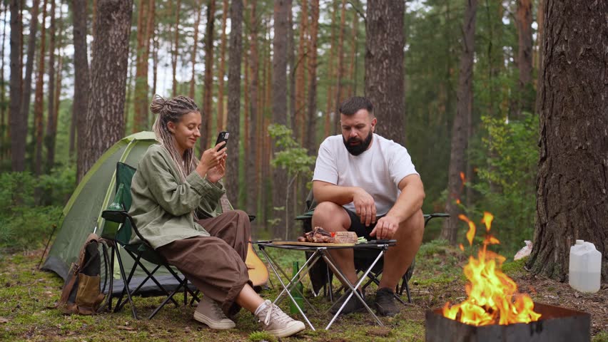 Beautiful young couple enjoying a romantic picnic in the woods, with a woman using her smartphone while a man prepares food near a crackling bonfire and their tent during a camping adventure