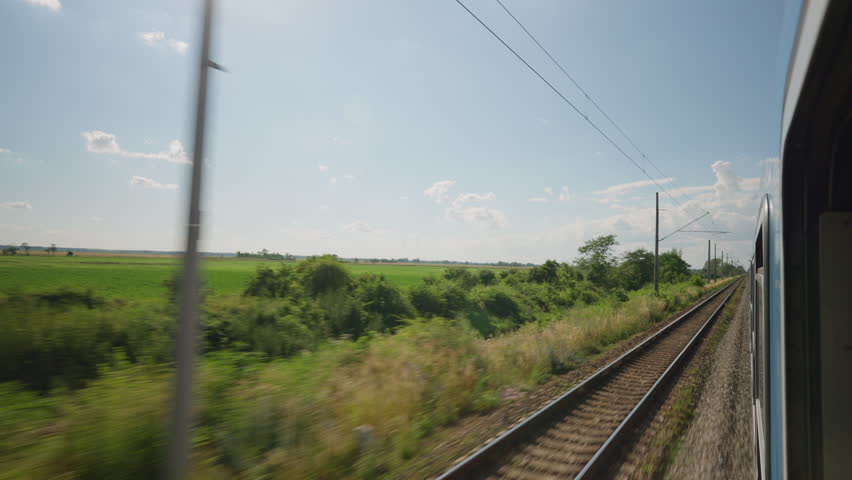 Point of view shot from a moving train showing railway tracks and green countryside on a sunny day. The rural landscape passes by quickly