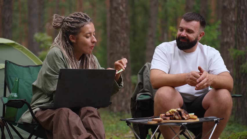 Young couple camping in the woods, with the woman working as a digital nomad on her laptop while the man relaxes, drinks beer, and cooks meat and vegetables on a portable barbecue grill