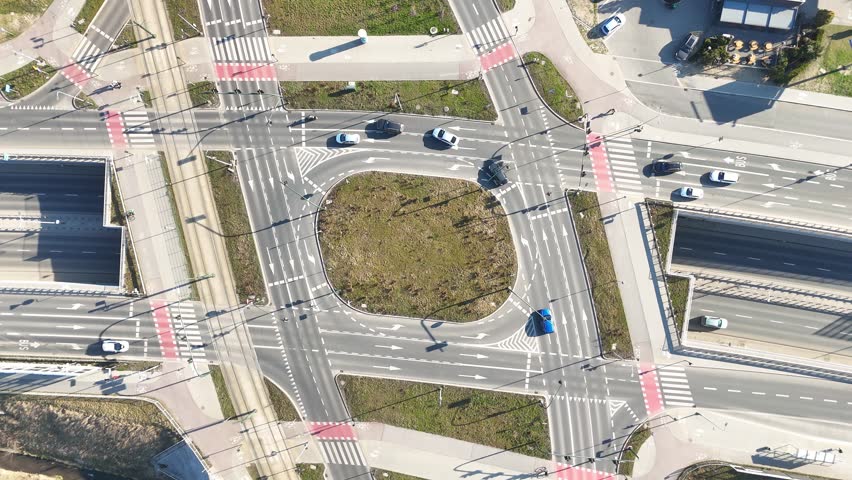 Aerial view of busy urban intersection with multiple roads, crosswalks, and vehicles navigating through traffic, showcasing pedestrians and surrounding green spaces in a city environment