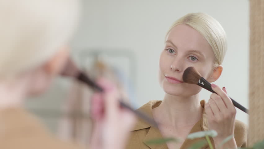 Fresh woman applying makeup with a soft brush during a beauty routine in front of a mirror. Young female, lady or model with natural skin and blonde hair creating a clean cosmetic look in a bright