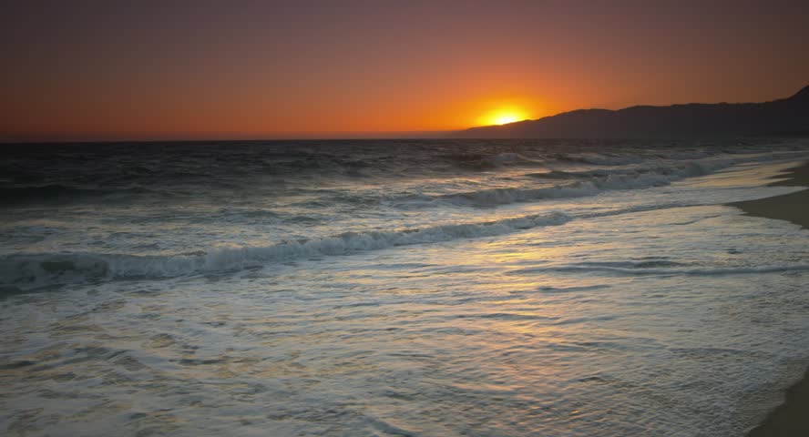 This 4K video shows a sunset over the Pacific Ocean at a beach in California, likely Santa Monica. The visual documents the sun on the horizon, orange light reflecting on the water, and white-foamed waves on the sand. The composition illustrates the coastline and silhouette hills, documenting North American maritime geography for nature and editorial media.