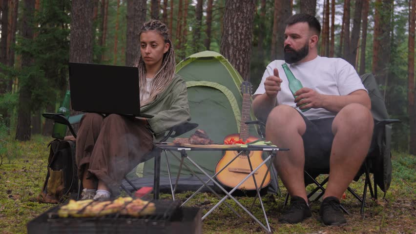 Young couple spending their vacation camping in a pine forest, with a digital nomad woman working on her laptop while her bored boyfriend sits beside her as meat grills on the barbecue