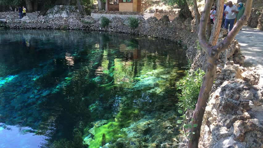 A stunning natural spring with turquoise and deep blue water, surrounded by stone walls and trees. The transparent water reveals rocks and aquatic vegetation beneath the surface, creating a serene and picturesque atmosphere. Visitors stand near the edge, admiring the beauty of this scenic nature spot. Perfect for themes of travel, nature, and outdoor exploration.
